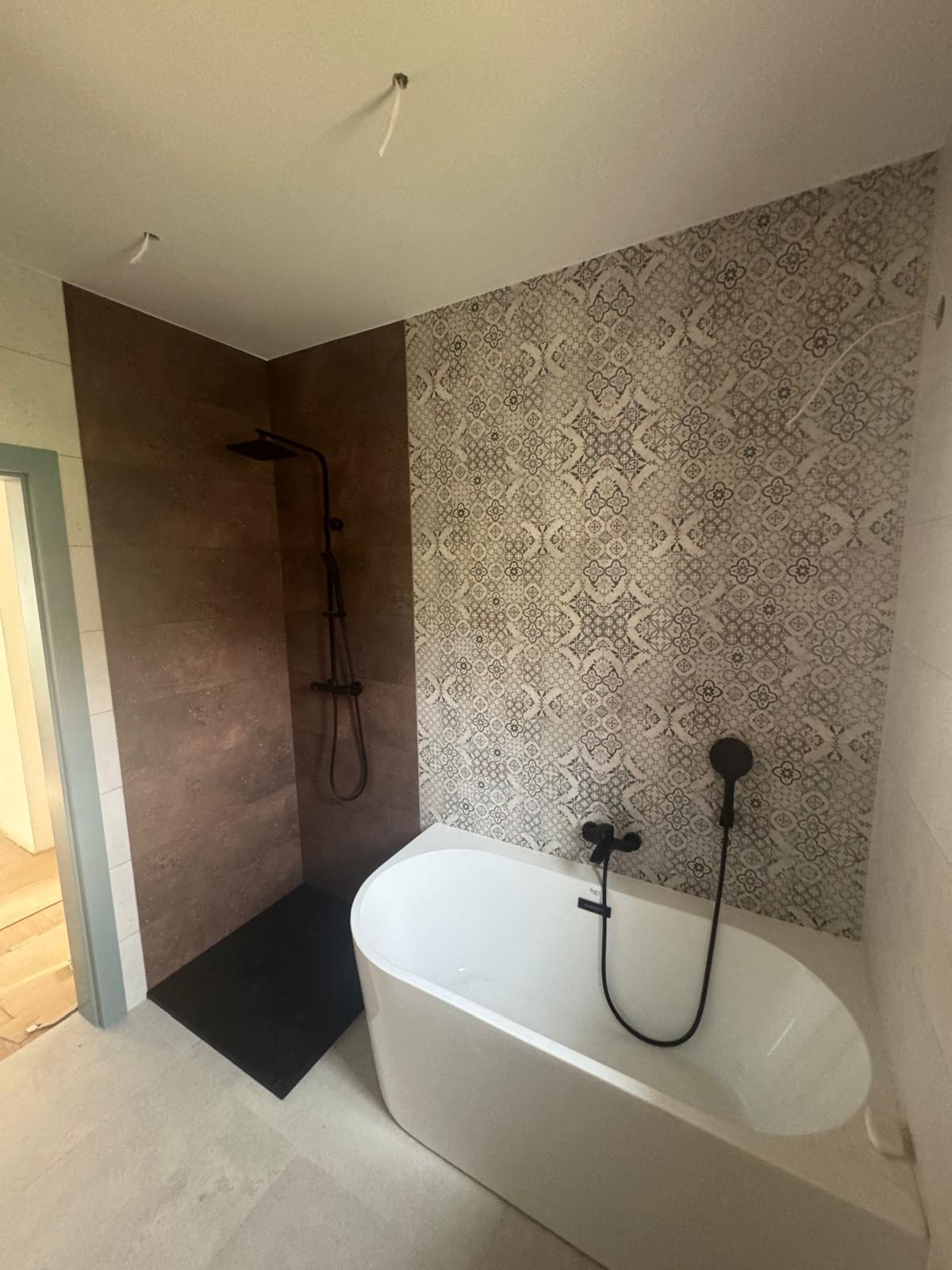 Nowoczesna łazienka po remoncie: biała wanna wolnostojąca, czarna armatura, płytki dekoracyjne na ścianie i brązowe płytki w strefie prysznicowej z odpływem liniowym.