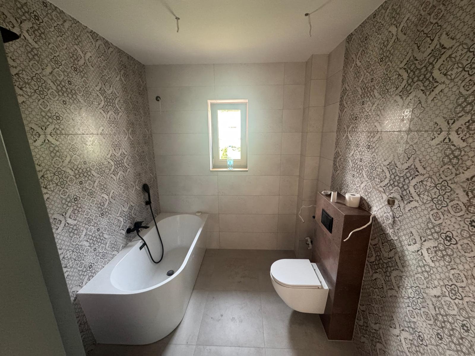 Wykończona łazienka z wanną, podwieszaną toaletą i wzorzystymi płytkami na ścianach. Widoczne niedokończone elementy oświetlenia.