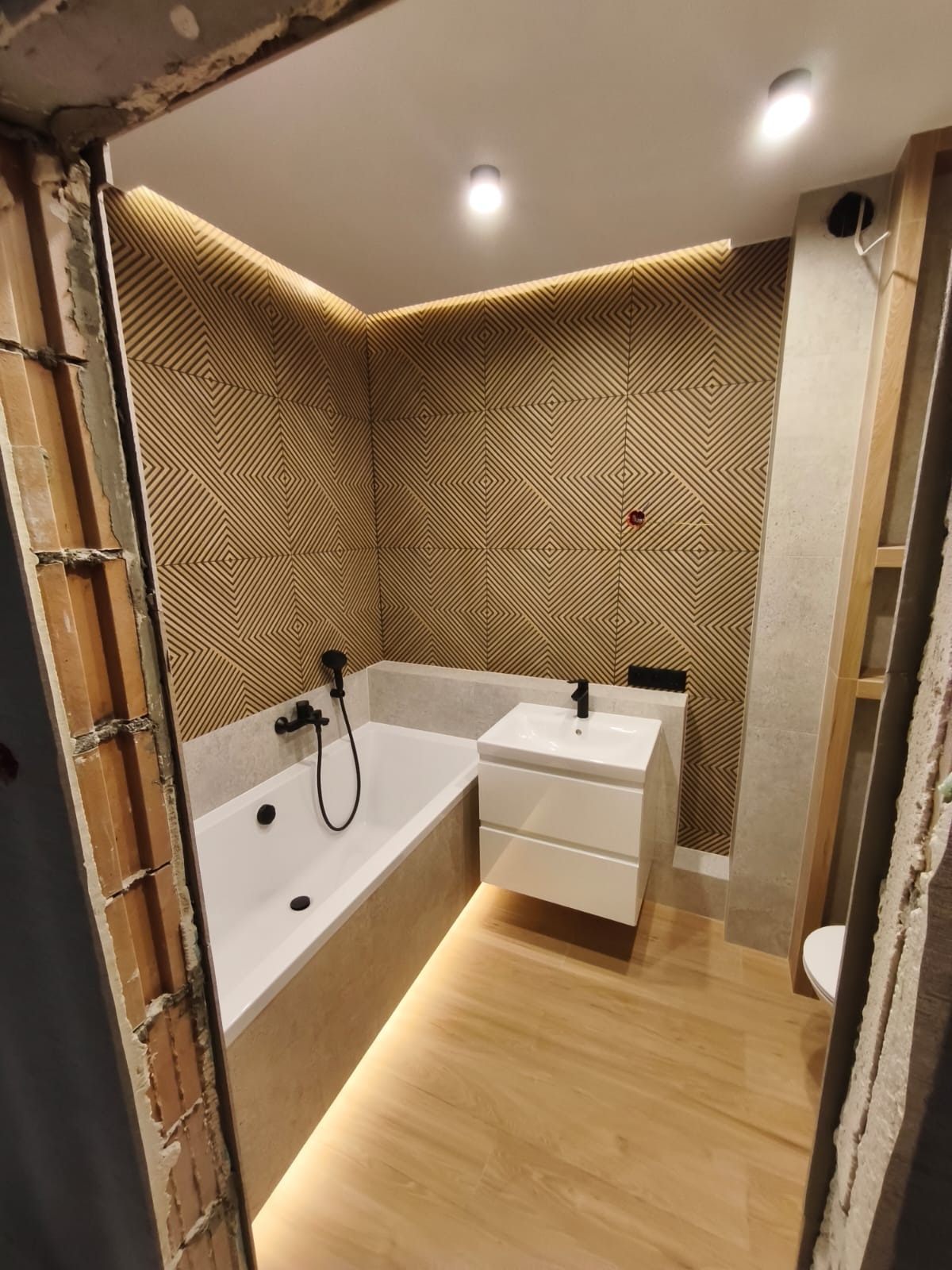 Nowoczesna łazienka po remoncie: biała wanna z czarną armaturą, umywalka z szafką, drewnopodobne płytki i dekoracyjna ściana z podświetleniem LED.