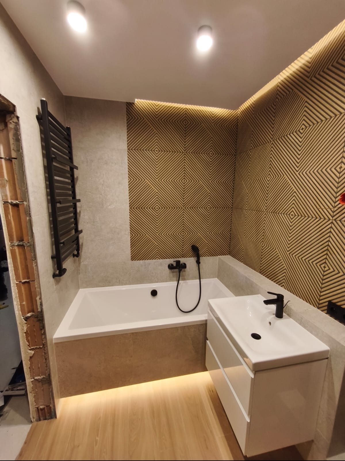 Nowoczesna łazienka z geometryczną ścianą, białą wanną i szafką umywalkową. Podświetlenie LED dodaje ciepła. Widoczny fragment niedokończonego wykończenia.