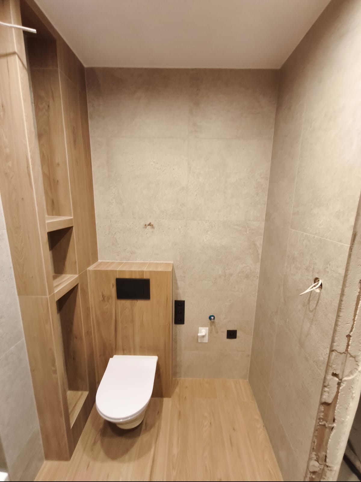 Nowoczesna łazienka z podwieszaną toaletą, drewnopodobne płytki na podłodze i ścianach, zabudowa z półkami. Minimalistyczny design, ciepłe odcienie.