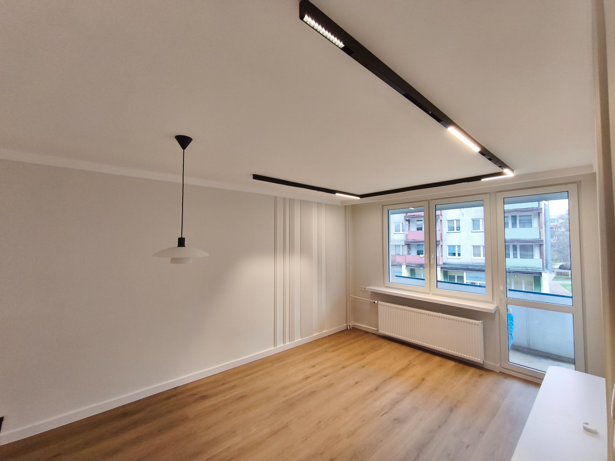 Jasne wnętrze po remoncie: panele podłogowe, białe ściany z dekoracyjnymi listwami, nowoczesne oświetlenie sufitowe i okno z widokiem na zewnątrz.