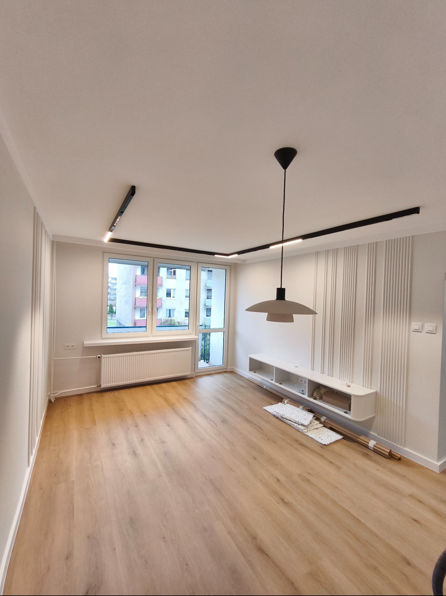 Jasne wnętrze po remoncie: panele podłogowe, białe ściany z dekoracyjnymi listwami, nowoczesne oświetlenie szynowe i wisząca lampa. Widok na okno i balkon.