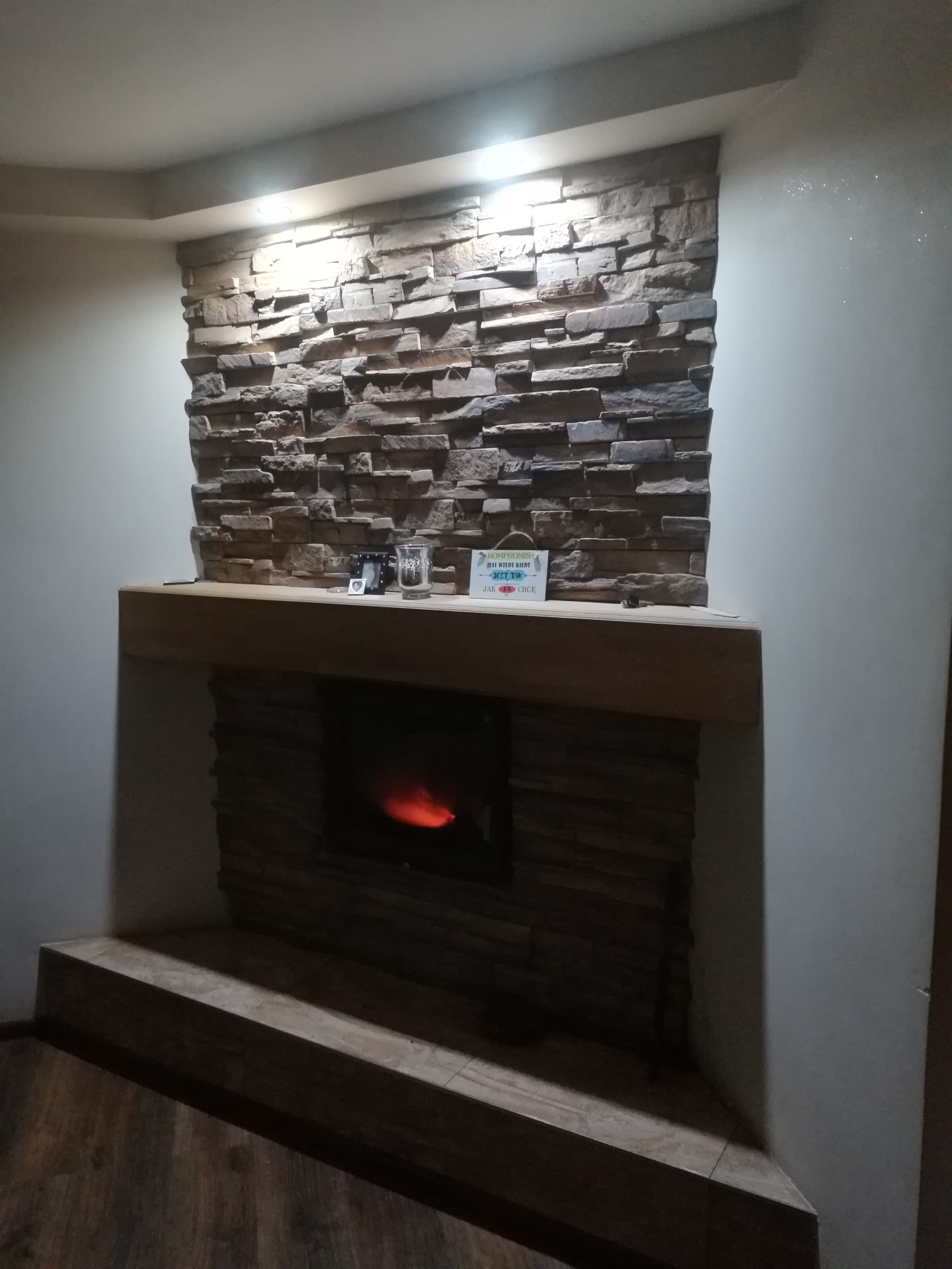 Komin obłożony dekoracyjnym kamieniem w odcieniach brązu, z drewnianym blatem, na którym stoją ozdoby, oraz widocznym paleniskiem z płomieniami, oświetlony ciepłym światłem.