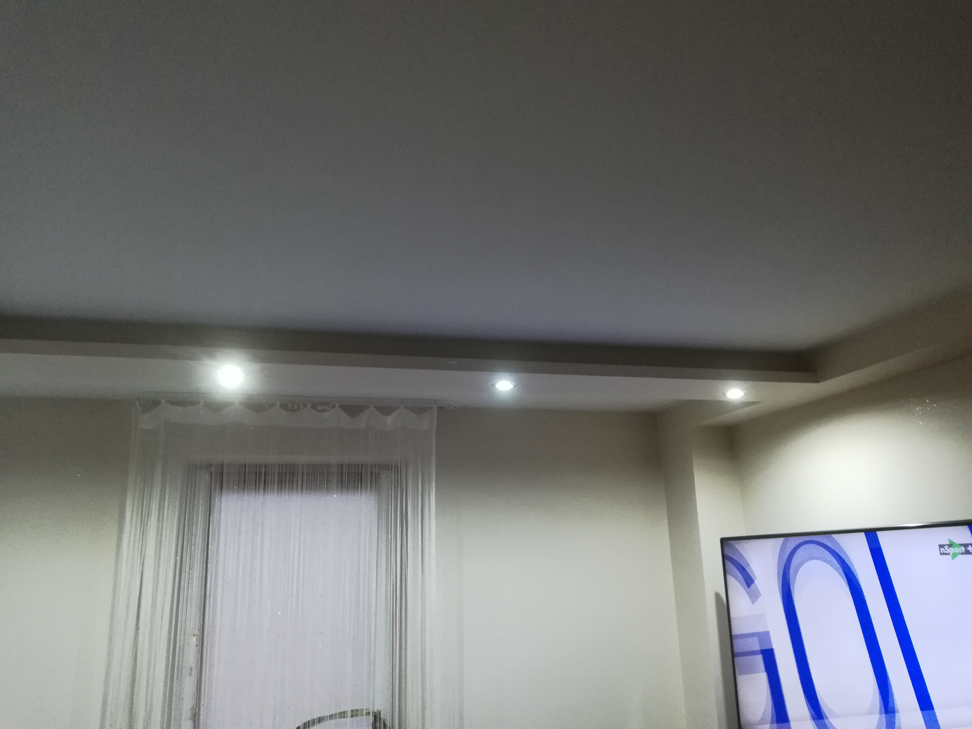 Podwieszany sufit z trzema punktowymi lampami LED w salonie, widok na okno z delikatnymi firankami i fragment telewizora.