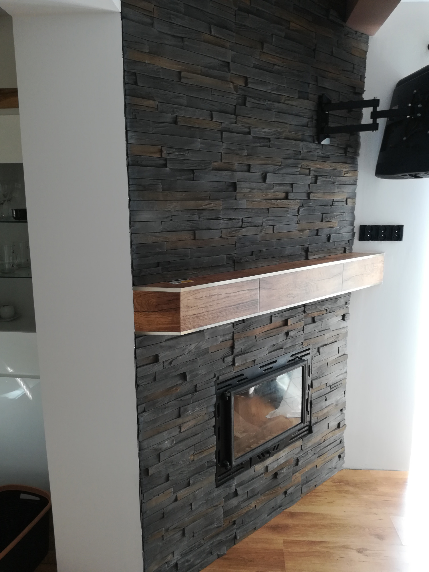 Ściana z kamienia dekoracyjnego z wbudowanym kominkiem i drewnianą półką, na ścianie zamontowany uchwyt do telewizora, widoczne gniazdka elektryczne.