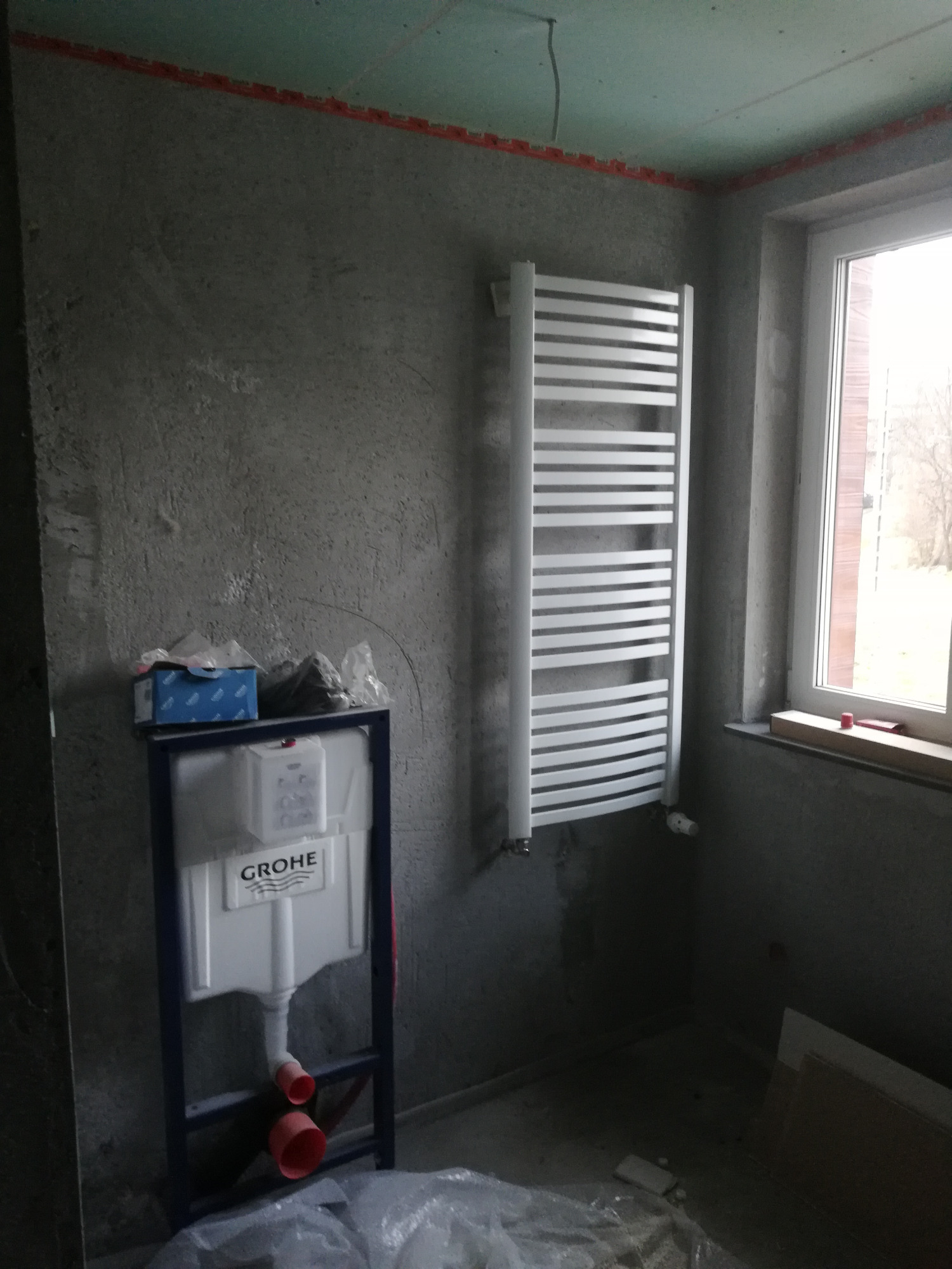 Wnętrze łazienki w trakcie remontu: widoczny stelaż podtynkowy WC marki Grohe, grzejnik drabinkowy na ścianie, surowy tynk i okno z widokiem na drzewa.