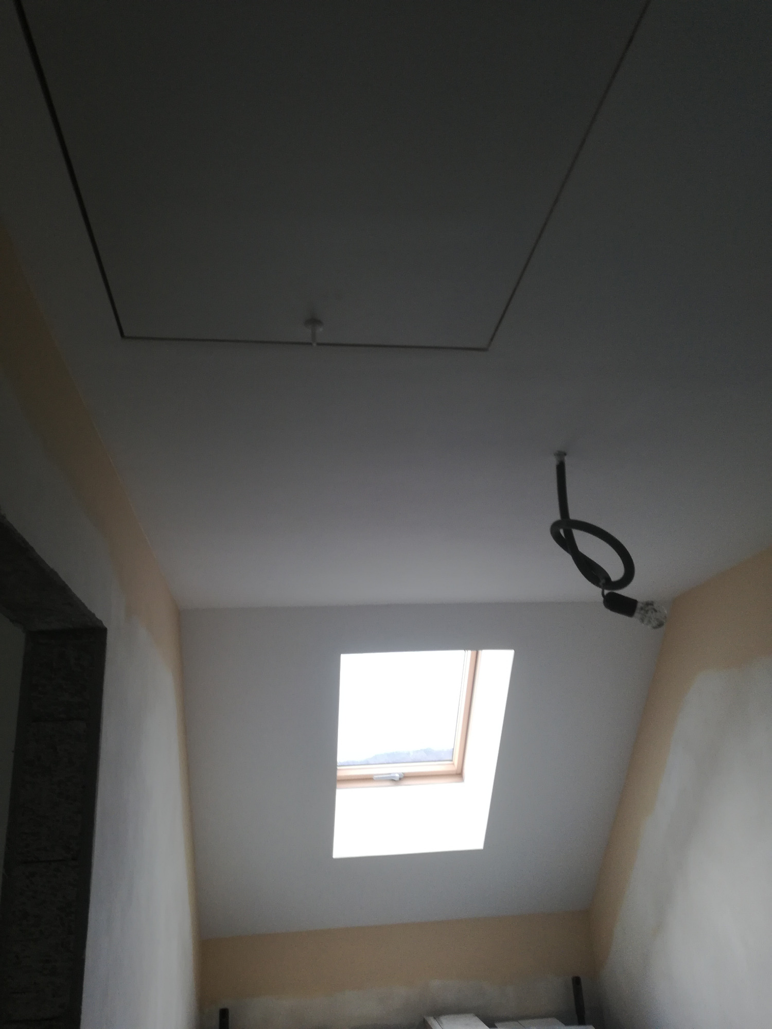 Poddasze w trakcie prac wykończeniowych: widoczne okno dachowe, częściowo pomalowane ściany i sufit, oraz przewód z żarówką zwisający z sufitu.