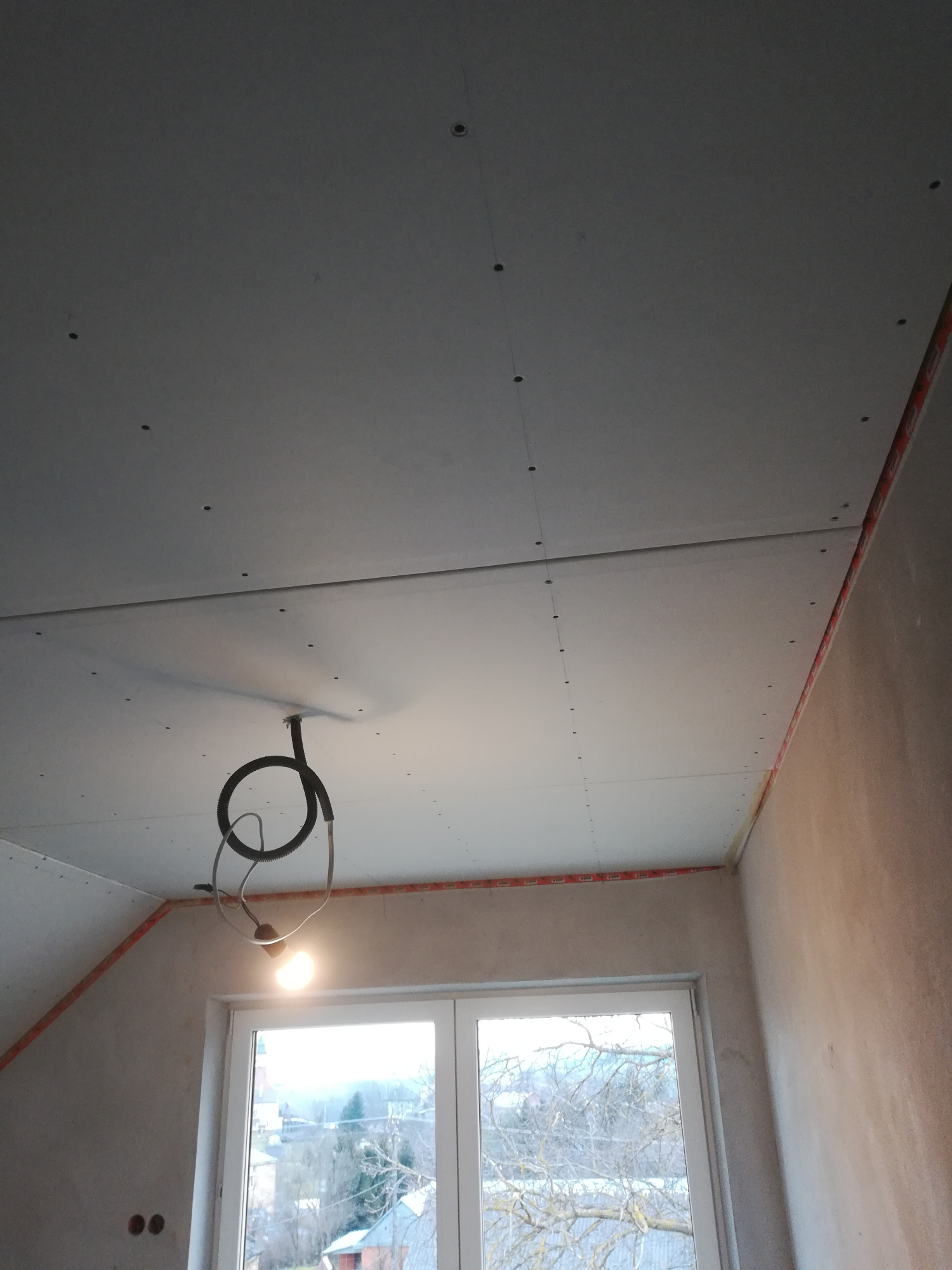 Montaż płyt gipsowo-kartonowych na poddaszu, widoczne łączenia, w tle okno z widokiem na drzewa.