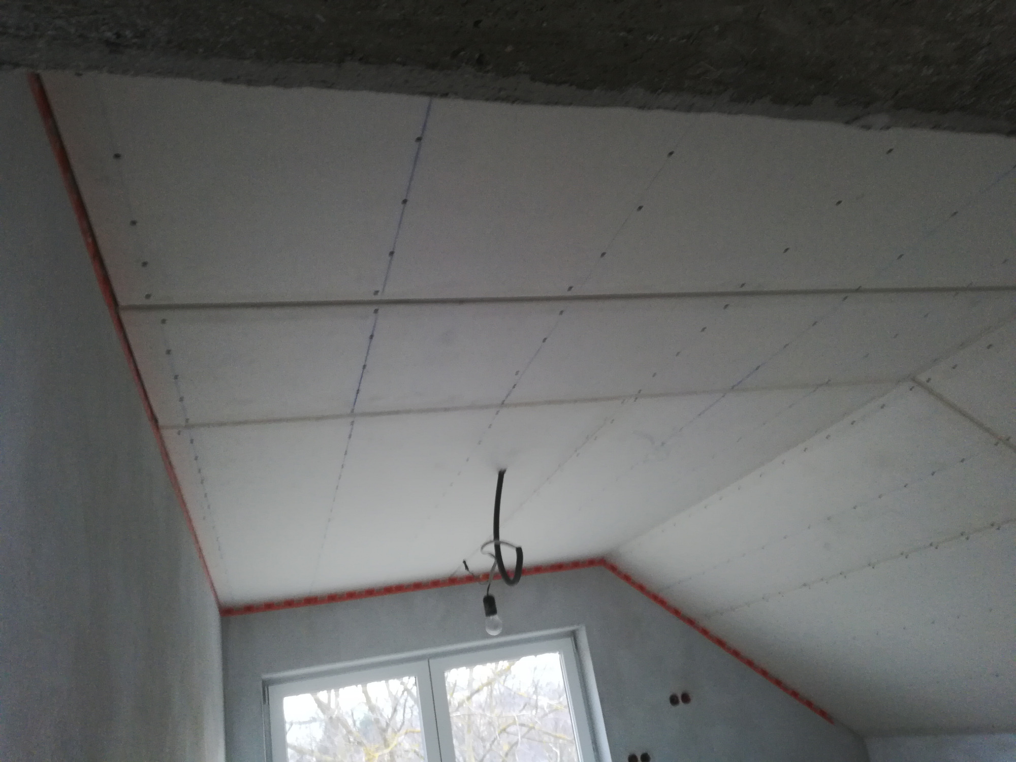 Montaż płyt gipsowo-kartonowych na poddaszu, widoczne profile i śruby, surowy stan wykończenia wnętrza, okno z widokiem na drzewa.