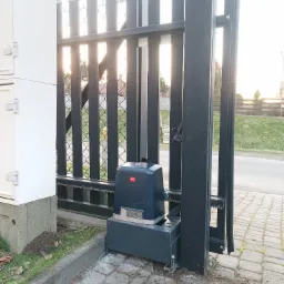 Szara, metalowa brama przesuwna z zamontowanym napędem automatycznym marki BFT, widoczne zębatki i skrzynka sterownicza obok bramy.