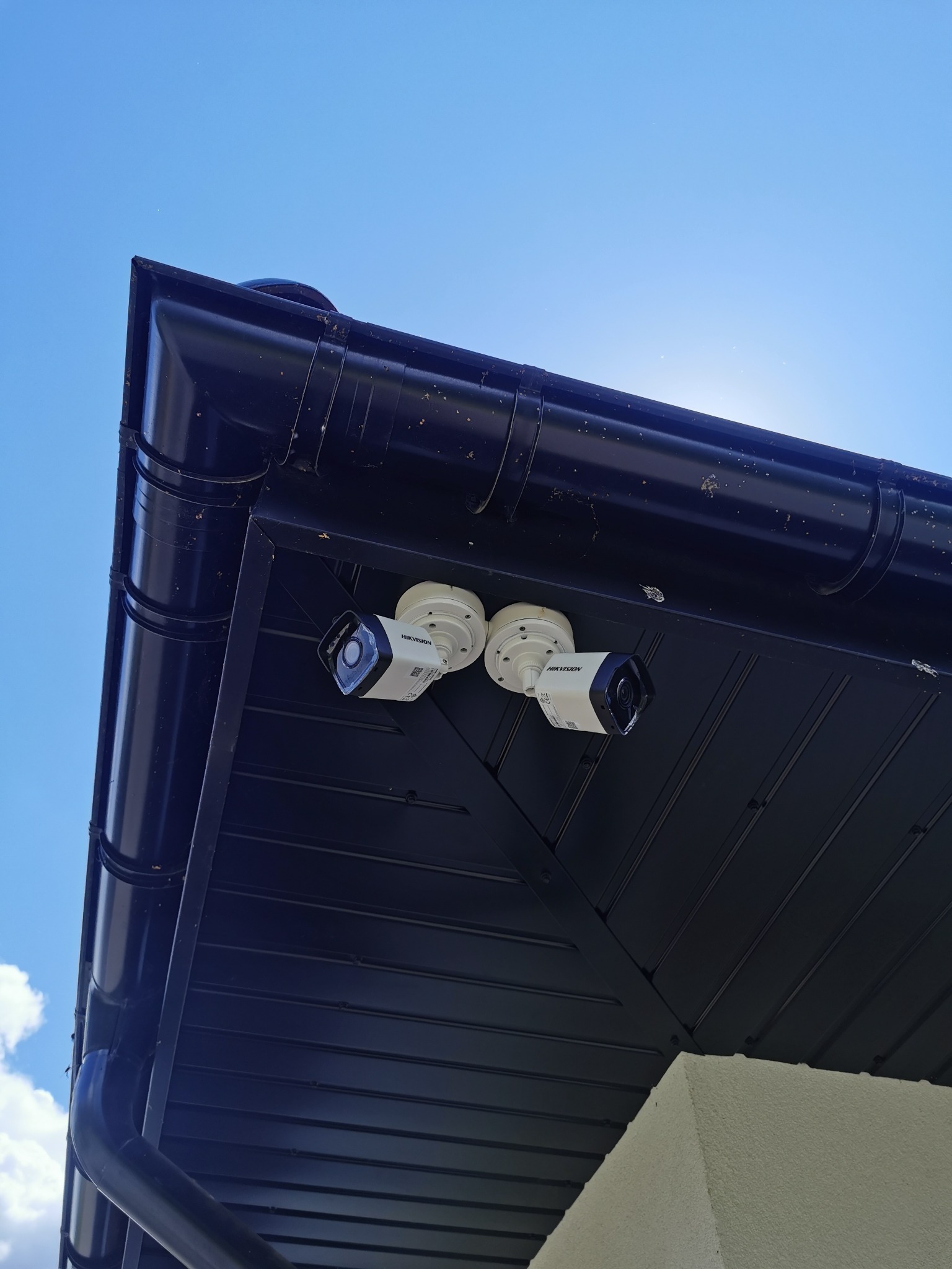 Dwie kamery monitoringu Hikvision zamontowane pod okapem dachu, widok z dołu na tle jasnego nieba.