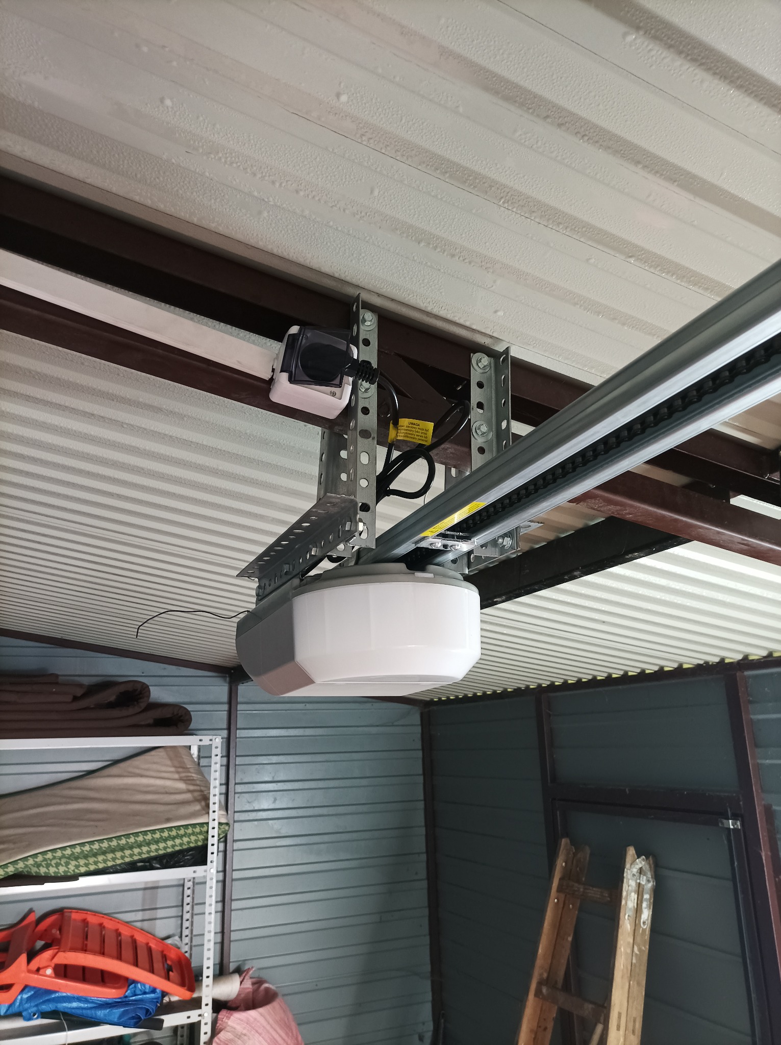 Zamontowany napęd bramy garażowej z widoczną szyną prowadzącą łańcuch, czujnikiem bezpieczeństwa i metalową konstrukcją mocującą do sufitu blaszanego garażu.