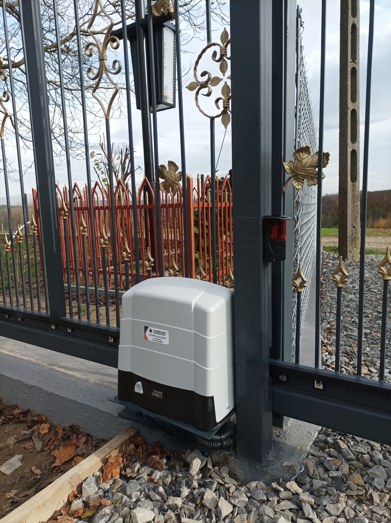 Szara brama wjazdowa z automatyką, ozdobnymi elementami w kolorze złota i lampą, widoczne liście na ziemi.