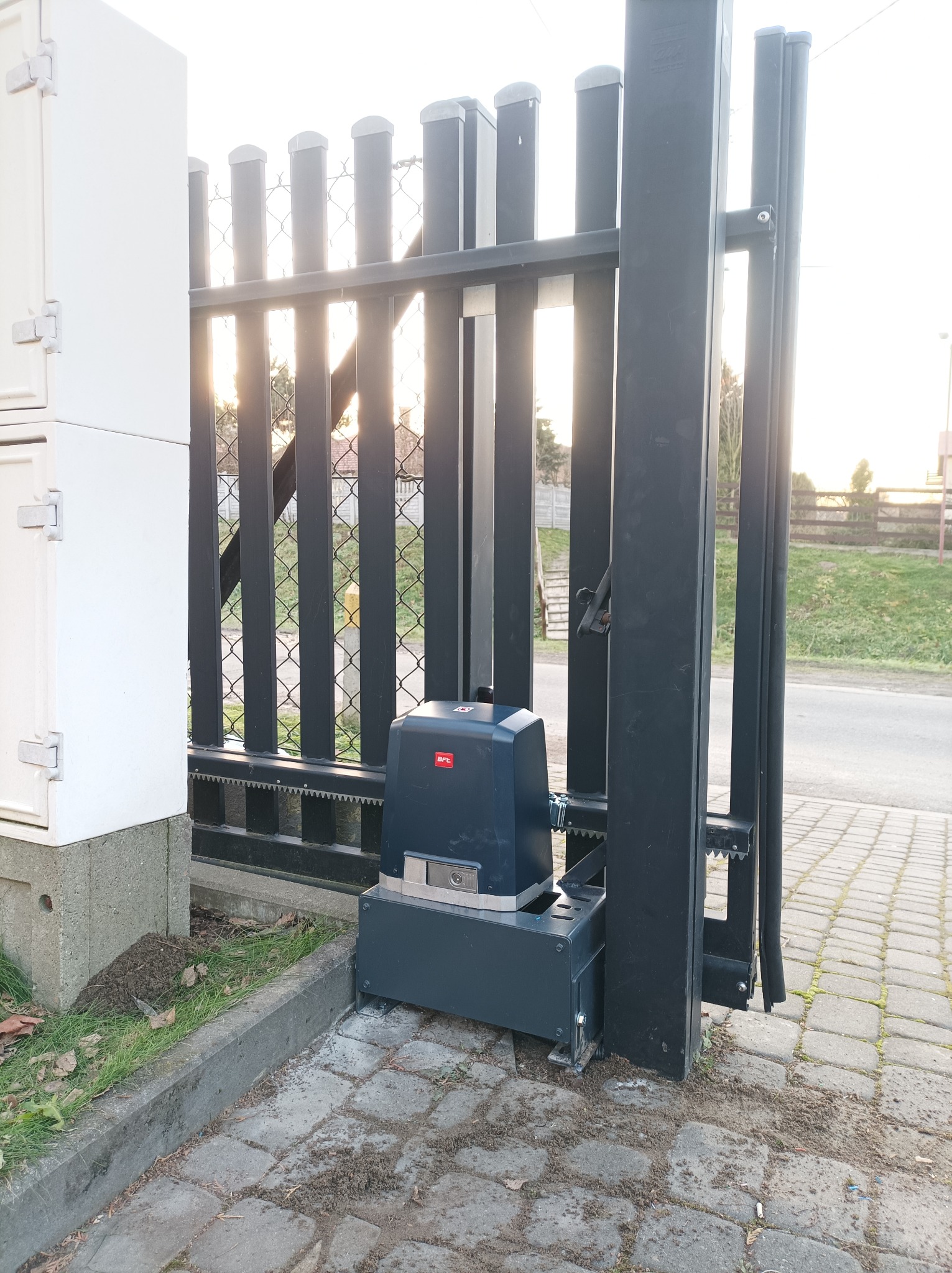 Szara, metalowa brama przesuwna z zamontowanym napędem automatycznym marki BFT, widoczne zębatki i skrzynka sterownicza obok bramy.