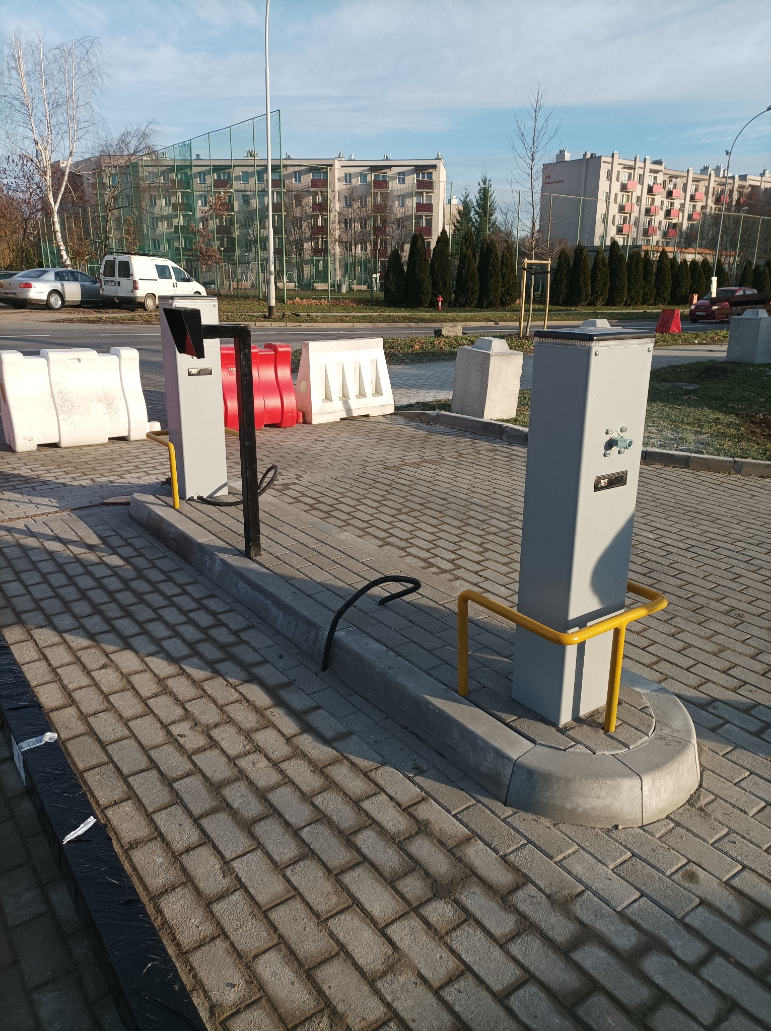 Dwa szare słupki z żółtymi barierkami na kostce brukowej, prawdopodobnie elementy systemu kontroli dostępu lub parkingu, z budynkami mieszkalnymi w tle.