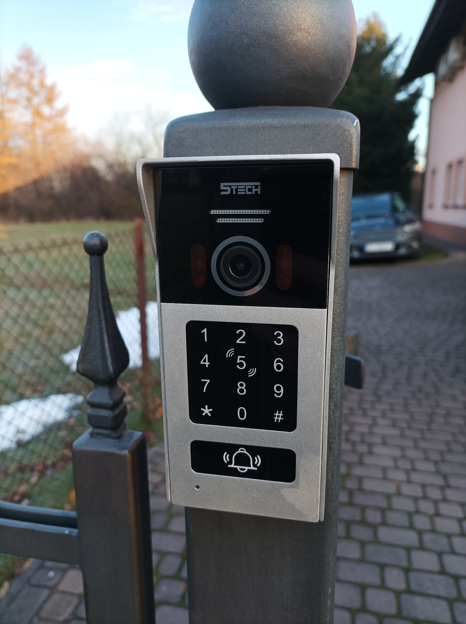 Zewnętrzny panel domofonowy z kamerą i klawiaturą numeryczną, zamontowany na metalowym słupku bramy wjazdowej; widoczny fragment ogrodzenia i samochód w tle.