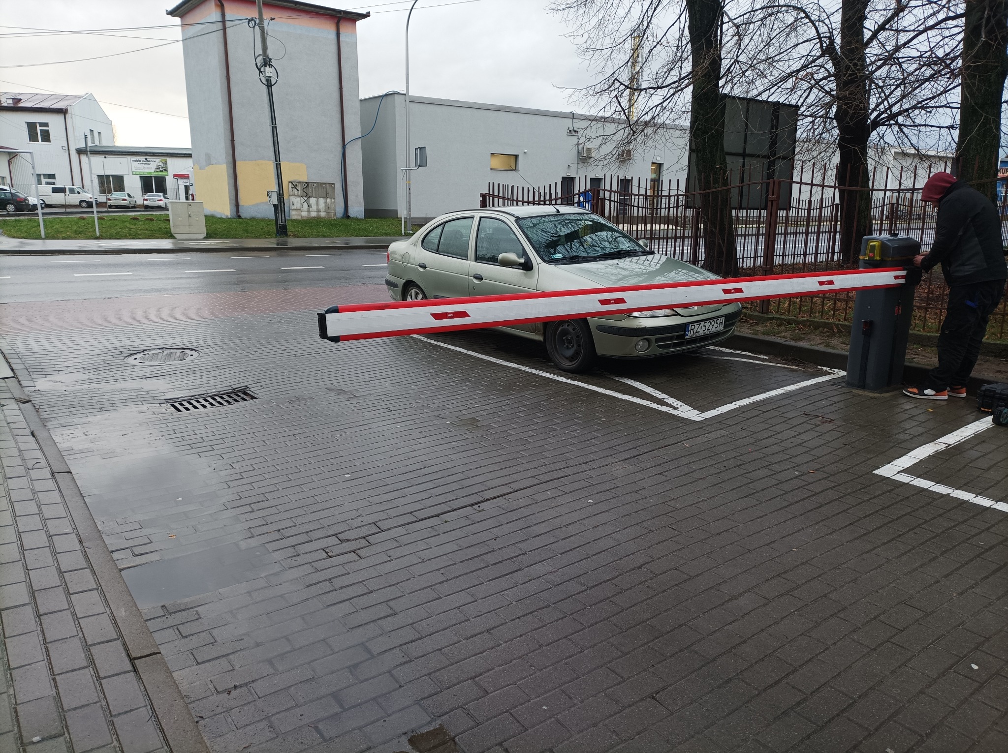 Szlaban z czerwoną i białą barierą opuszczony przed zaparkowanym srebrnym samochodem na mokrym brukowanym parkingu, osoba w ciemnej odzieży przy automacie szlabanu.