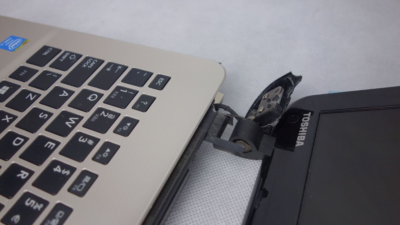 Uszkodzony laptop Toshiba z wyłamanym zawiasem ekranu, widoczny fragment klawiatury i obudowy ekranu na tle szarej powierzchni.
