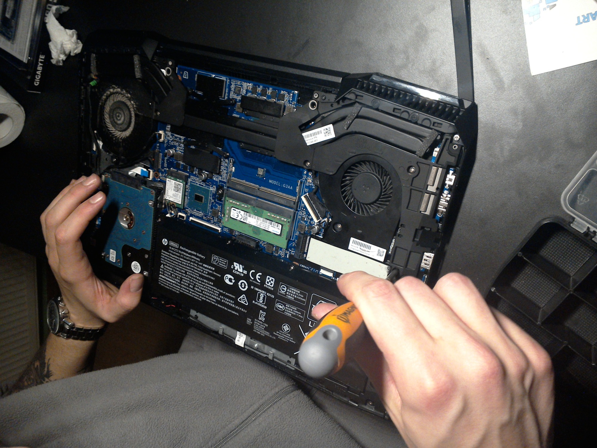 Otwarty laptop z widoczną płytą główną, wentylatorami i baterią, serwisant odkręca śrubkę pomarańczowym śrubokrętem, widoczny model G3AA, obok leżą chusteczki higieniczne i pudełko po Gigabyte.