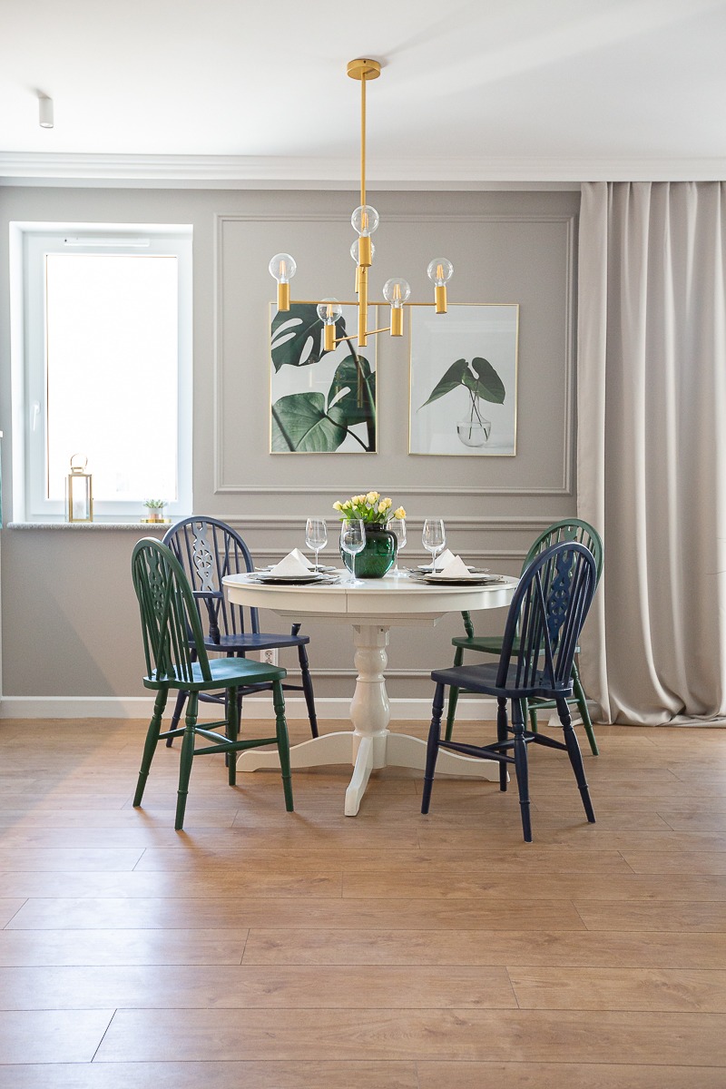 Jadalnia z okrągłym białym stołem, czterema różnokolorowymi krzesłami, złotym żyrandolem i dekoracjami roślinnymi na ścianie.