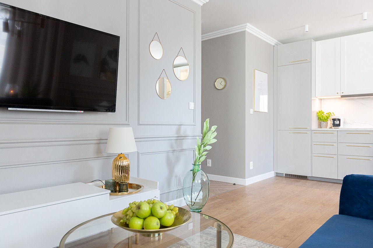 Eleganckie wnętrze mieszkania: szara ściana z telewizorem i trzema okrągłymi lusterkami, złota lampka na białej szafce RTV, szklany stolik z owocami i wazonem, w tle biała zabudowa kuchenna...