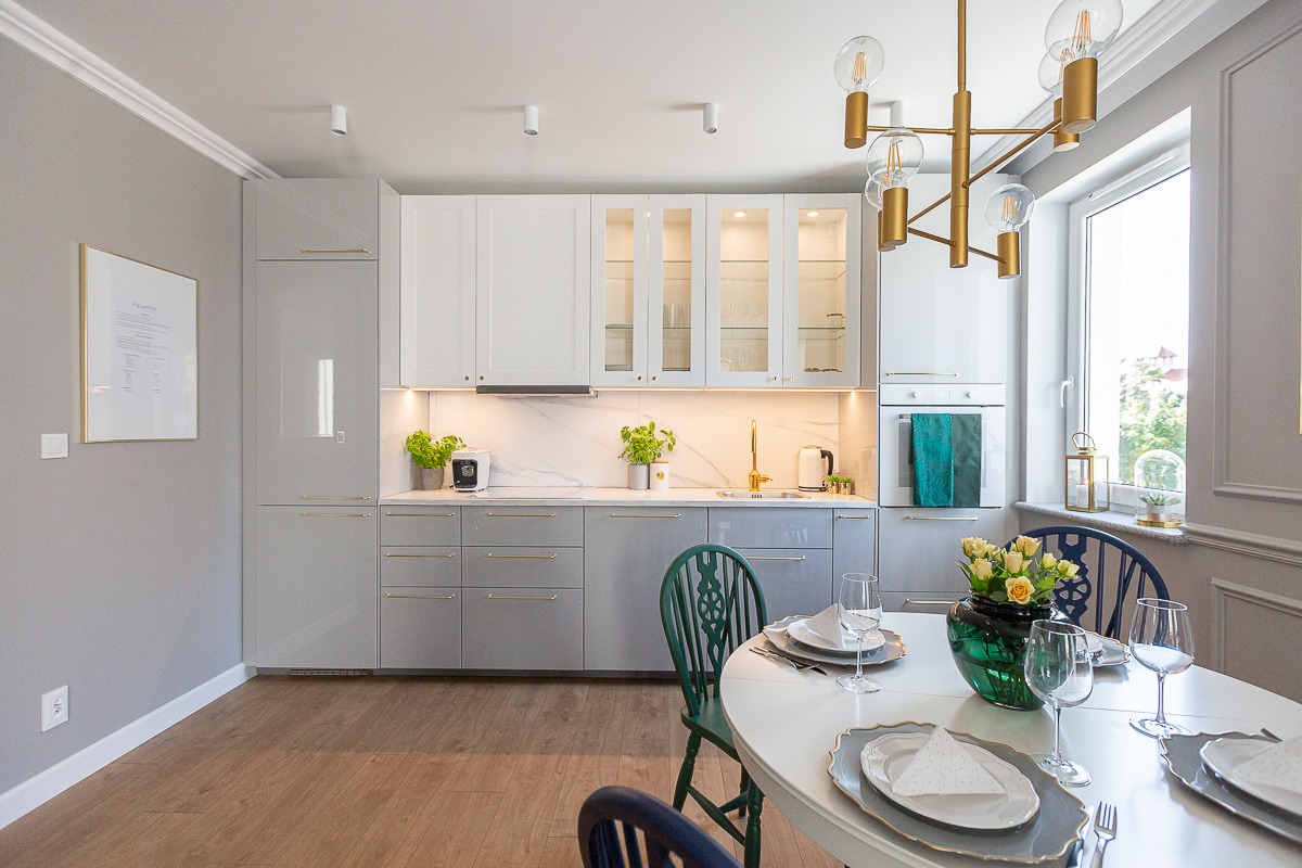 Nowoczesna kuchnia w szaro-białej tonacji z elementami złota: szafki z połyskiem, marmurowy backsplash, złota armatura i designerska lampa wisząca nad stołem z nakryciem.