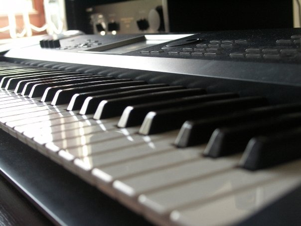 Czarno-białe klawisze syntezatora muzycznego uchwycone pod kątem, z widocznymi przyciskami i potencjometrami w tle, oświetlone naturalnym światłem wpadającym z okna.