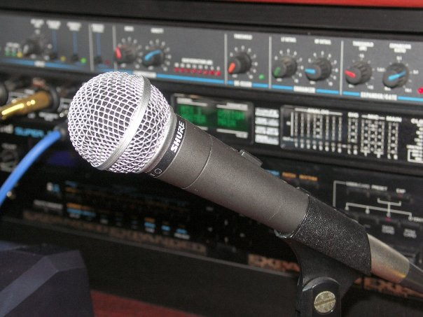 Mikrofon Shure na statywie w zbliżeniu, w tle sprzęt studyjny z pokrętłami i wyświetlaczami.