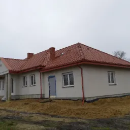 Nowy dom jednorodzinny z czerwoną dachówką, widoczne kominy z cegły i orynnowanie, teren wokół domu w trakcie prac ziemnych.