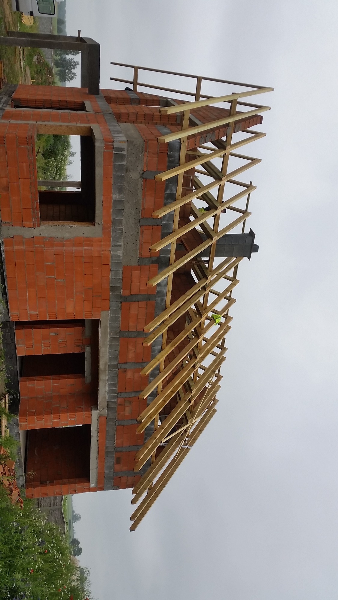 Surowa konstrukcja dachu z drewnianymi belkami na murowanym budynku z cegły, widok z dołu na pochmurnym tle.