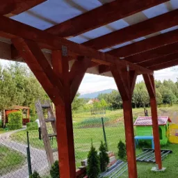 Zadaszenie tarasu z drewnianą konstrukcją i przezroczystym pokryciem, widok z wnętrza na ogród z trampoliną i domkiem dla dzieci.