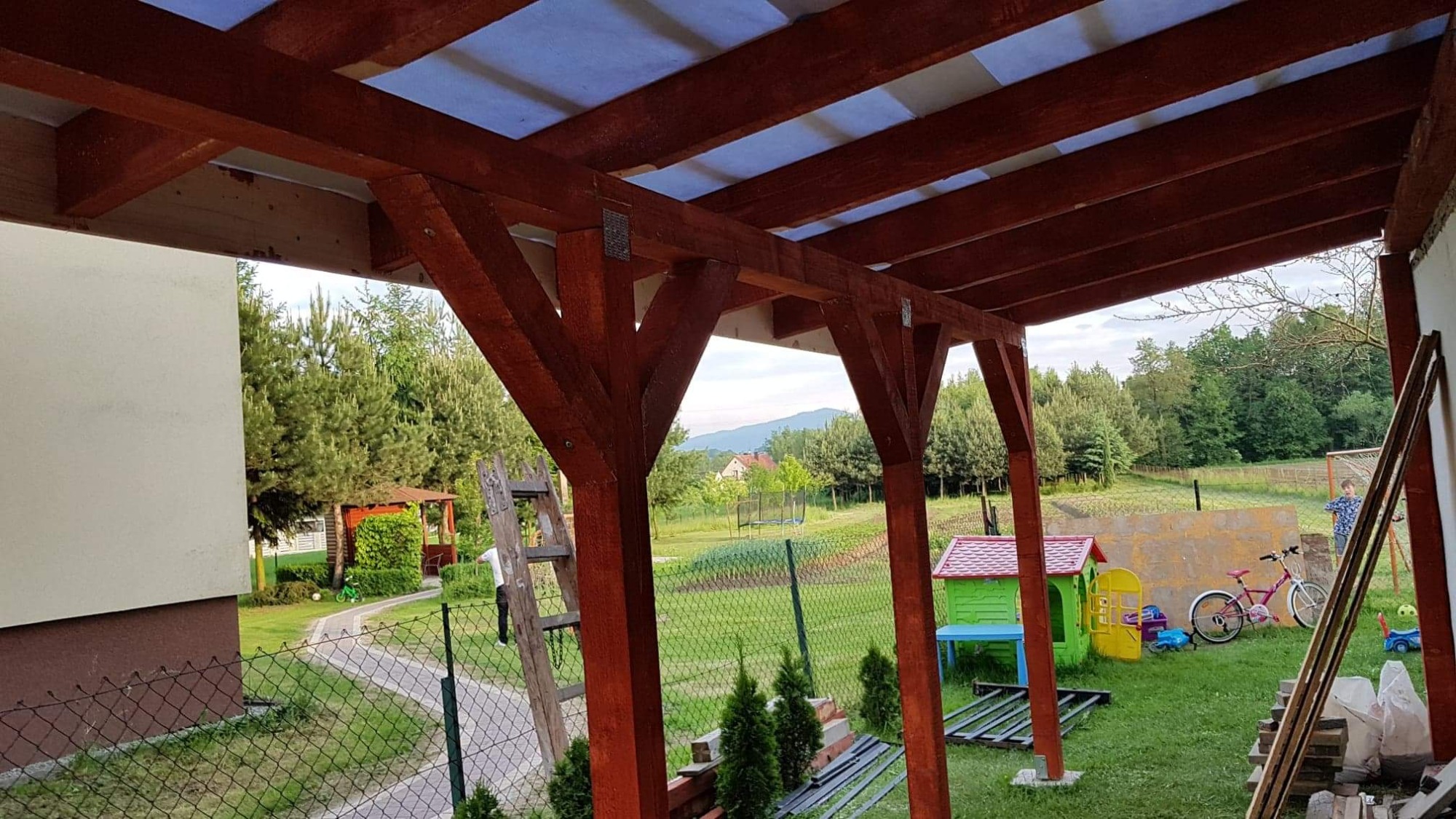 Zadaszenie tarasu z drewnianą konstrukcją i przezroczystym pokryciem, widok z wnętrza na ogród z trampoliną i domkiem dla dzieci.