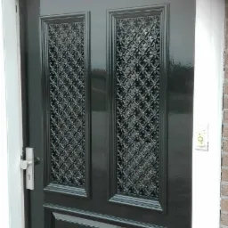 Błyszcząca, ciemnozielona, odnowiona stolarka drzwiowa z ozdobnymi, ażurowymi wstawkami, widoczna klamka i dzwonek.