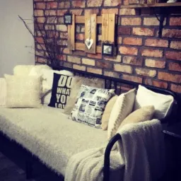 Rustykalna ściana z cegły z drewnianą dekoracją i poduszkami na metalowej sofie z miękkim kocem.