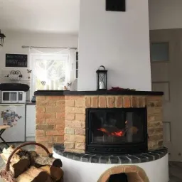 Ceglany kominek z paleniskiem, płomienie widoczne za szybą, kosz z drewnem obok, w tle kuchnia z lodówką i mikrofalówką.