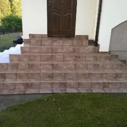 Wykończone schody wejściowe do domu, wyłożone brązowymi płytkami ceramicznymi, z widocznymi fugami i fragmentem trawnika oraz elewacji budynku.