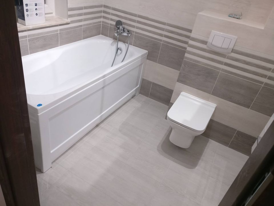 Nowoczesna łazienka z białą wanną, toaletą i szaro-białymi płytkami ułożonymi w poziome pasy, widok z perspektywy drzwi.