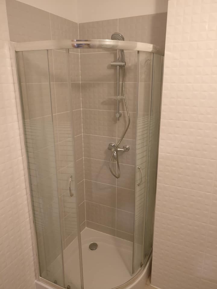 Narożna kabina prysznicowa z przesuwnymi drzwiami, wykończona beżowymi płytkami z delikatną fakturą, zamontowana w jasnej łazience.