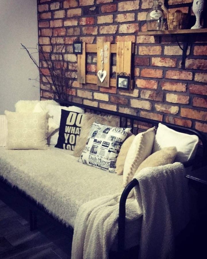 Rustykalna ściana z cegły z drewnianą dekoracją i poduszkami na metalowej sofie z miękkim kocem.