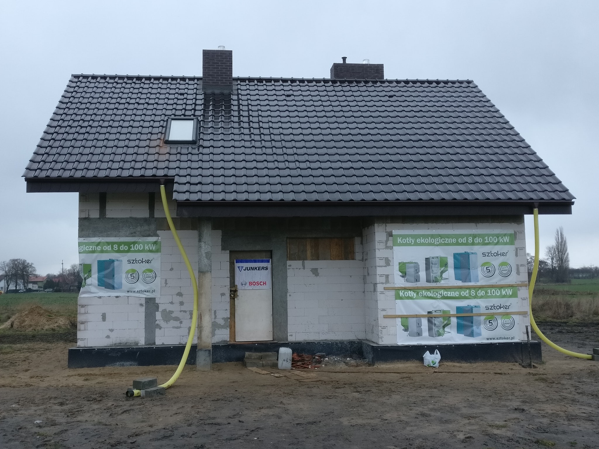 Surowy stan domu jednorodzinnego z ciemną dachówką, oknem dachowym i częściowo otynkowaną elewacją, na której wiszą banery reklamowe kotłów grzewczych, z widocznymi żółtymi rurami doprowadzającymi...