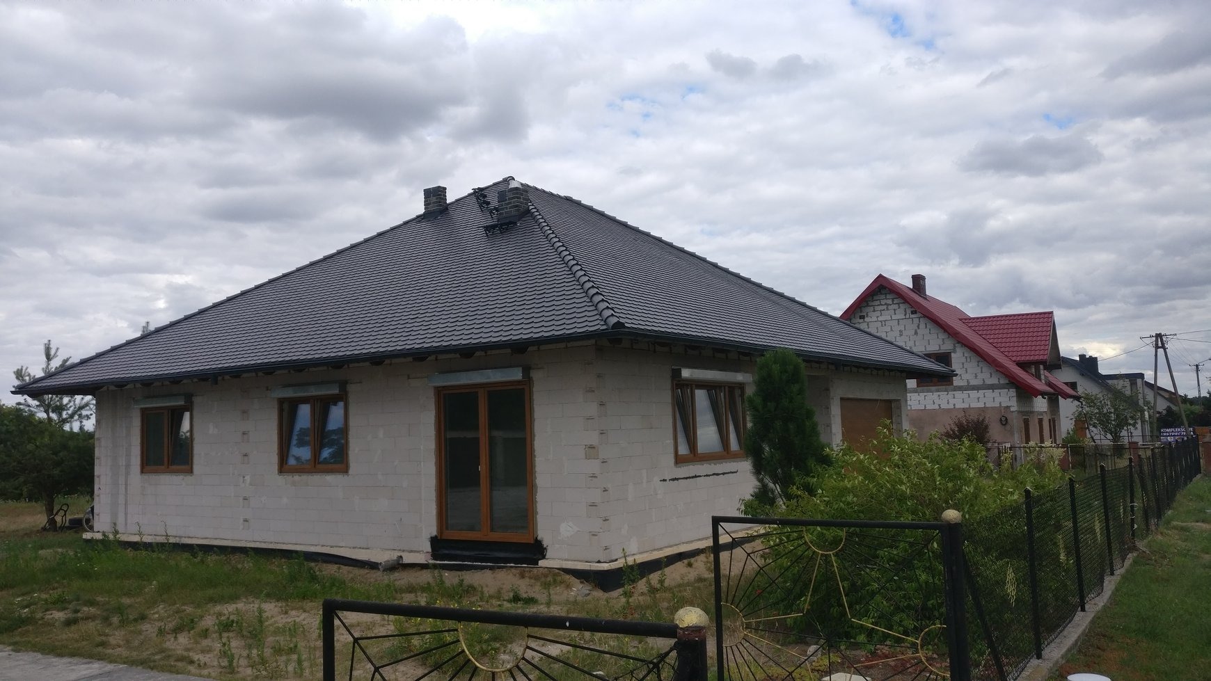 Nowy dom w stanie surowym zamkniętym z ciemną dachówką i drewnianymi oknami, widoczne niedokończone obróbki blacharskie komina i brakujące rynny.