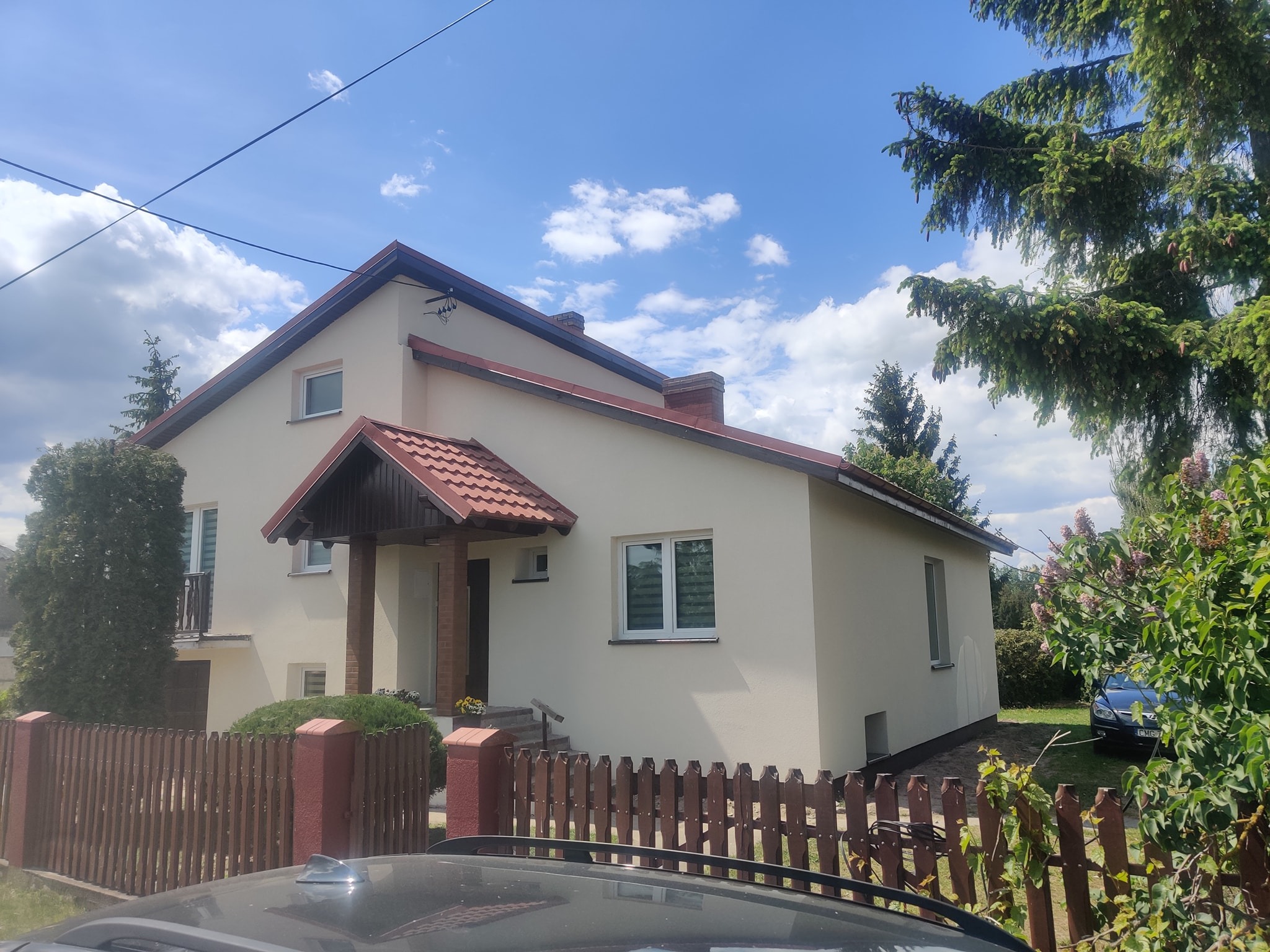 Otynkowany dom jednorodzinny z brązową dachówką i drewnianym ogrodzeniem, widok zza samochodu, błękitne niebo z chmurami w tle.