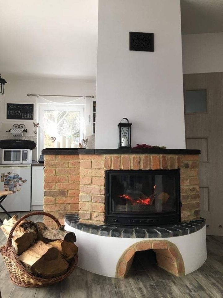 Ceglany kominek z paleniskiem, płomienie widoczne za szybą, kosz z drewnem obok, w tle kuchnia z lodówką i mikrofalówką.