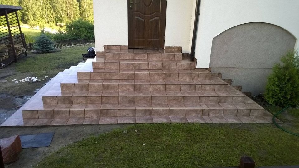 Wykończone schody wejściowe do domu, wyłożone brązowymi płytkami ceramicznymi, z widocznymi fugami i fragmentem trawnika oraz elewacji budynku.