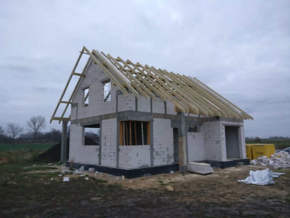 Surowy stan domu jednorodzinnego z białych bloczków, w trakcie budowy dachu z drewnianą konstrukcją, widoczne okna bez szyb, na zewnątrz sterta bloczków i ziemi.