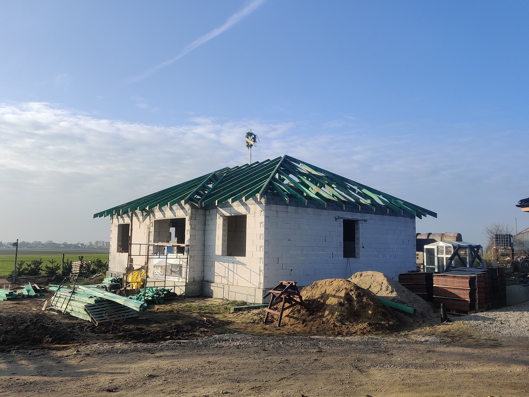 Budowa domu jednorodzinnego w stanie surowym otwartym, widoczne ściany z bloczków, więźba dachowa z zielonymi łatami, stos materiałów budowlanych i sterta piasku na placu budowy.