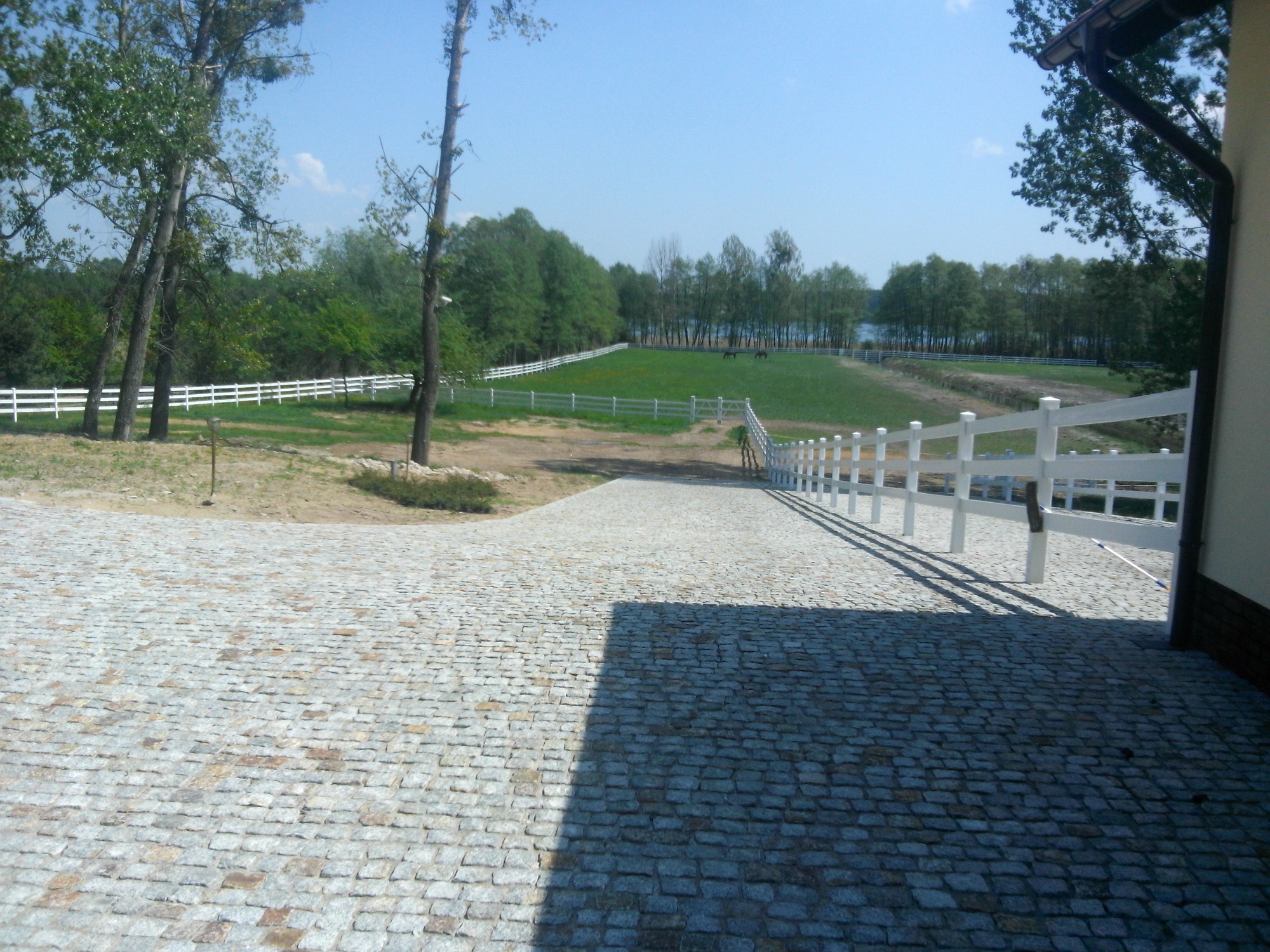 Szeroki podjazd z kostki granitowej prowadzący do posesji z białym ogrodzeniem farmerskim, w tle zielony krajobraz z drzewami i jeziorem.