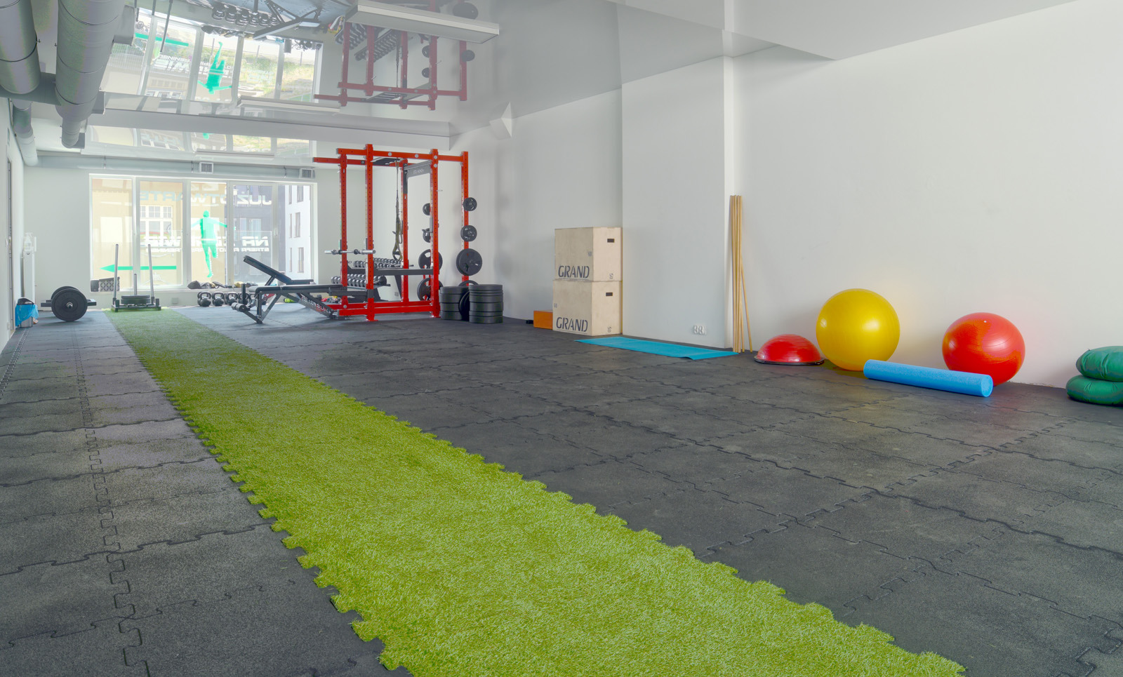 Jasne, przestronne wnętrze siłowni z zielonym pasem na podłodze, sprzętem do ćwiczeń siłowych, skrzyniami plyometrycznymi i piłkami gimnastycznymi.