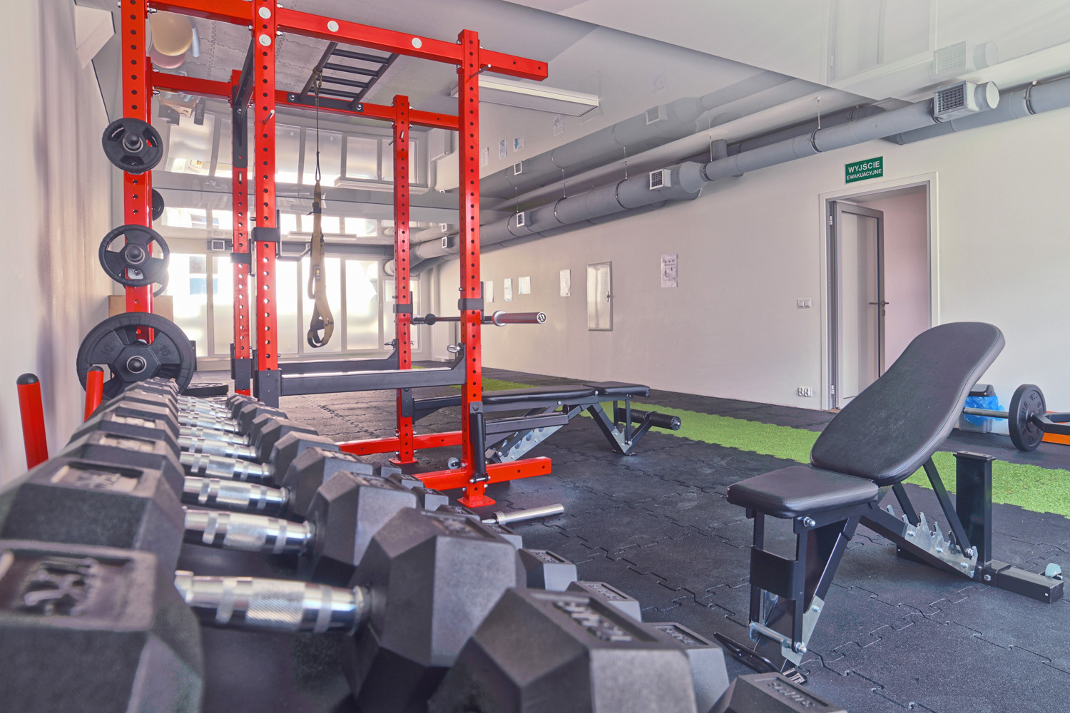 Wnętrze siłowni z rzędem hantli, czerwonym stojakiem do ćwiczeń i ławkami, widok na okno i drzwi ewakuacyjne
